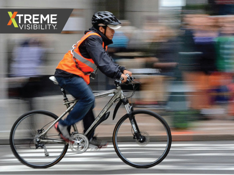 A man wearing an orange safety vest riding a bike through city streets.